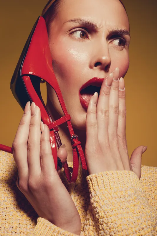 Surprised woman with red lipstick holds red high heel shoe near face, wearing yellow sweater. Concept for fashion, beauty, and unexpected emotions in studio with warm lighting.