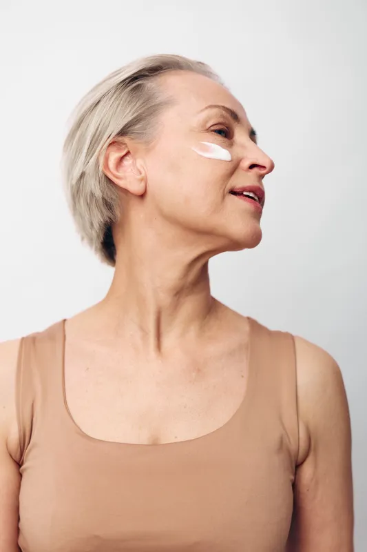 Mature woman with short grey hair applies white facial cream on cheek. Natural skincare concept with close-up profile view against plain white background for beauty and self-care.