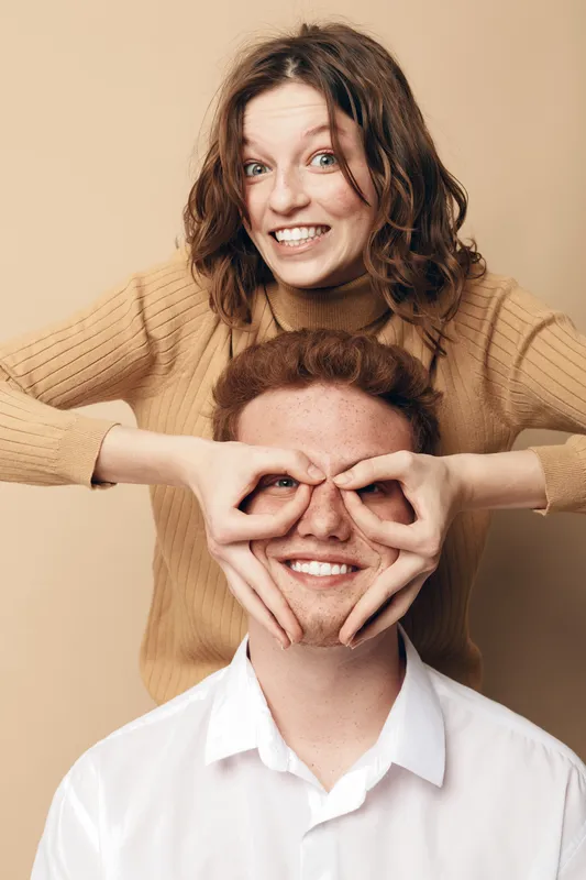 Playful young woman in brown sweater makes glasses gesture with hands on smiling man in white shirt face. Studio portrait on beige background. Concept of friendship and fun moments.