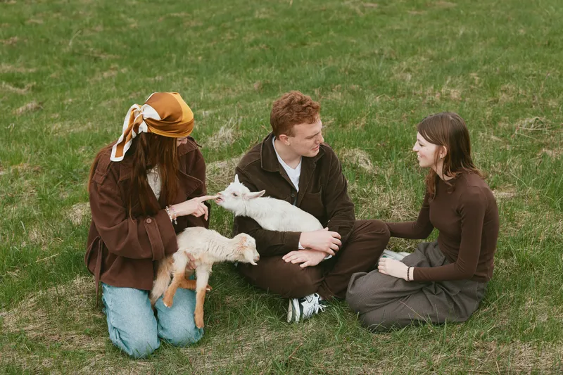 Lifestyle scene with three friends on a grassy field, softly stroking a young goat as it nuzzles a palm, warm smiles and gentle interaction creating a wholesome park setting