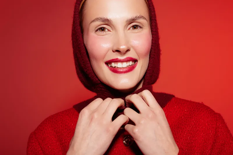 Smiling woman in red sweater and hood looks happy on red background. Close-up portrait with natural makeup and bright lipstick. Concept of joy, fashion, and winter style.
