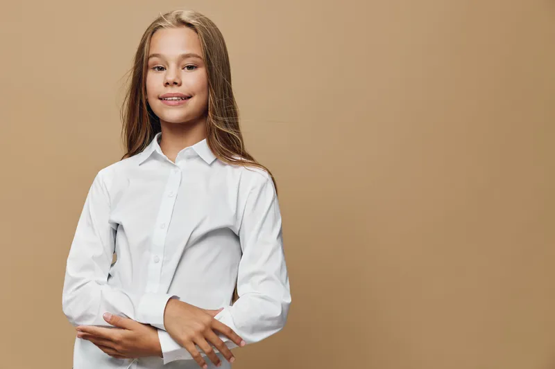 Girl child with long hair wearing white shirt smiling and posing in front of beige background with confidence and happiness. Child model portrait with casual elegant clothing, youth and beauty.