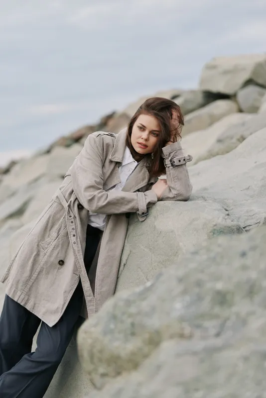 Young woman in beige knitted coat leans on large rocks outdoors. Overcast sky creates moody atmosphere. Casual fashion, autumn style, thoughtful expression in natural setting.
