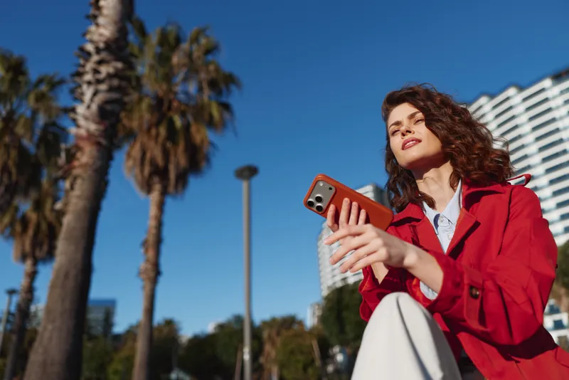 Young woman in red coat holds smartphone outdoors with palm trees and modern buildings on sunny day, looking up with confident expression.