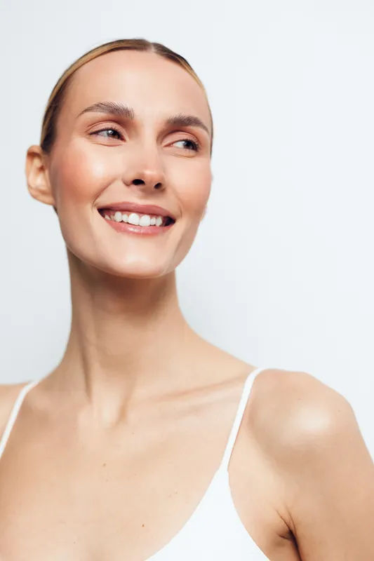 Smiling woman with clear skin looks to side, natural light highlights her face, minimal makeup, bare shoulders, simple white background, fresh and confident expression