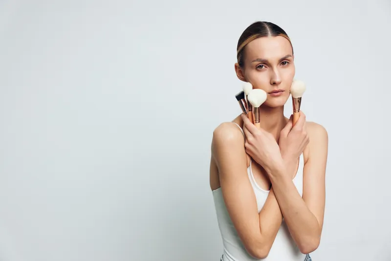 Woman applying makeup with a brush, focusing on her face, beauty in a studio, grooming concept, calm minimalist portrait, white background, soft light