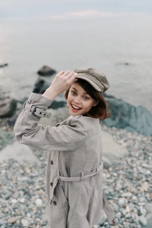 Smiling young woman in beige coat and hat poses on rocky beach with ocean background. Natural light highlights joyful expression and casual autumn fashion style outdoors.