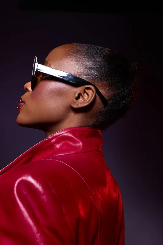 Profile of a confident black woman with dark skin wearing black sunglasses and a red leather jacket. Studio portrait with dramatic lighting on a dark purple background. Fashion concept.