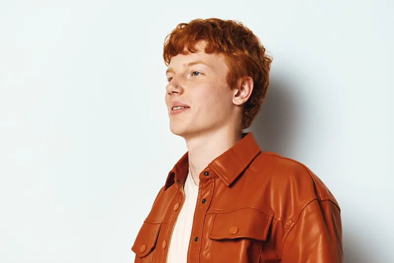 Young man with red hair wearing brown leather jacket standing against white background looking aside with confident expression in studio portrait.