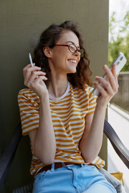 A relaxed woman sits on a balcony wearing a striped t shirt and jeans, smiling while talking on her phone, with a cigarette in her other hand. A casual home lifestyle moment.