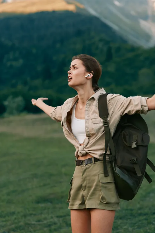 Young woman with backpack standing outdoors with arms outstretched enjoying nature. Concept of lifestyle, freedom, hiking, and adventure in a green mountain landscape.