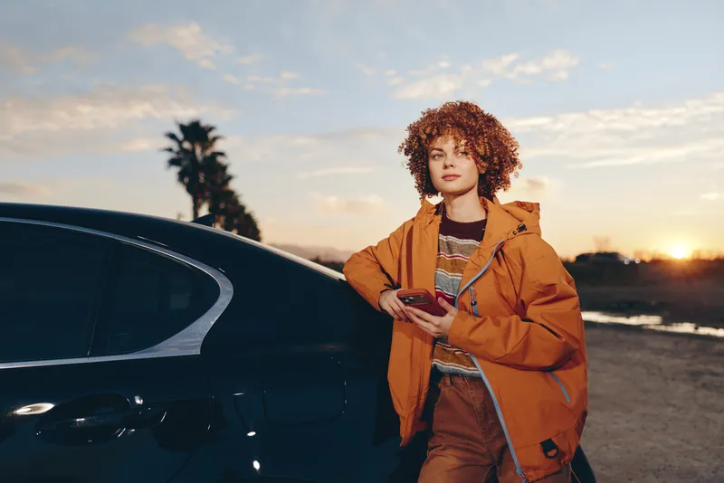 Woman lifestyle car outdoors sunset jacket curly hair portrait road, young woman stands beside parked car on a coastal road, brown jacket, holding small bag, warm evening light, confident smile
