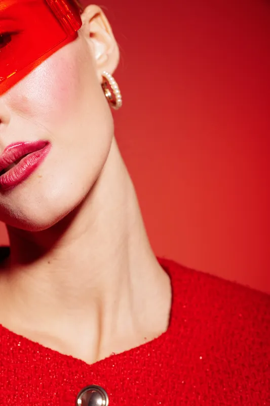 Close-up of a woman wearing red lipstick and a red outfit with a shiny red hat partially covering her face. The vibrant red background enhances the bold fashion and makeup concept.