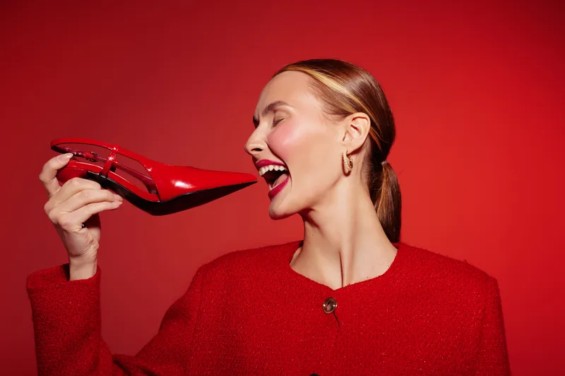 Smiling woman in red jacket holds red high heel shoe near face on red background. Concept of fashion, style, glamour, and bold color contrast in studio lighting.