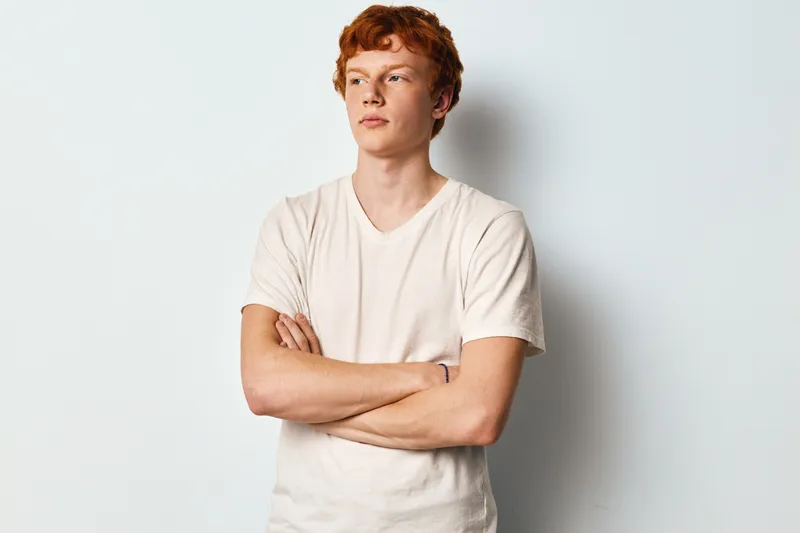 Young man with red hair and light skin wearing plain white t shirt standing with crossed arms looking sideways on white background. Casual style, confident facial expression.