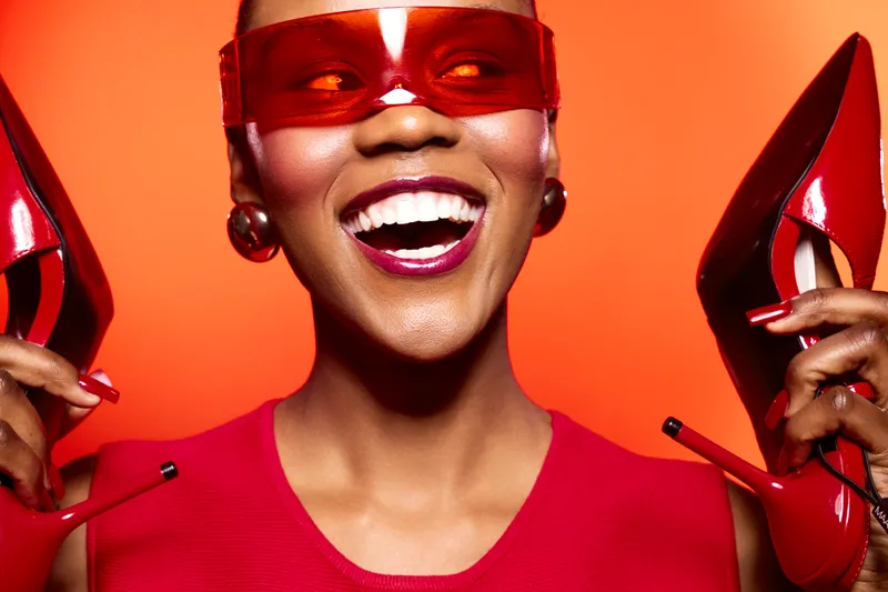 Smiling black woman with dark skin holds red high heels near her face wearing red dress and red sunglasses. Bright orange background highlights joyful fashion concept and stylish accessories.
