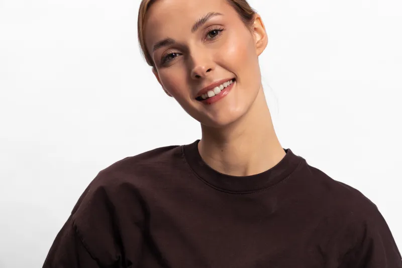 Smiling young woman with natural makeup wearing dark brown sweater looks at camera on white background. Portrait of happy female model with casual style and confident expression.