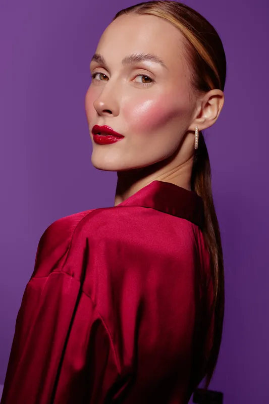 Portrait of a confident woman with glossy red lips and sleek hair wearing a red satin blouse against a purple background. Concept of beauty, fashion, elegance, and makeup.