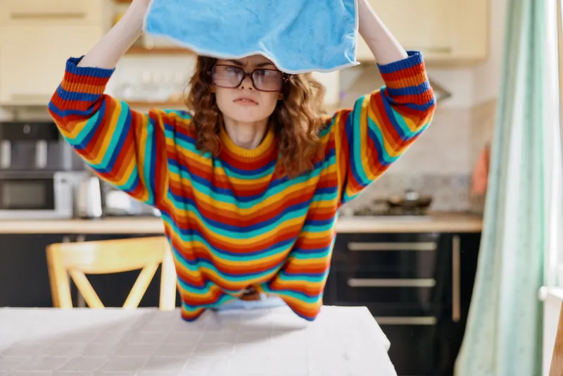 Young girl with curly hair and glasses lifts a blue towel over her head in a bright kitchen, turning a simple moment into playful color and curiosity.