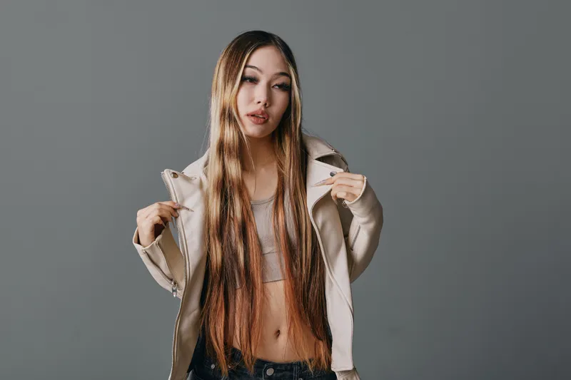 Young woman with long straight hair wearing casual jacket and crop top posing against gray background with confident expression and relaxed posture in studio.