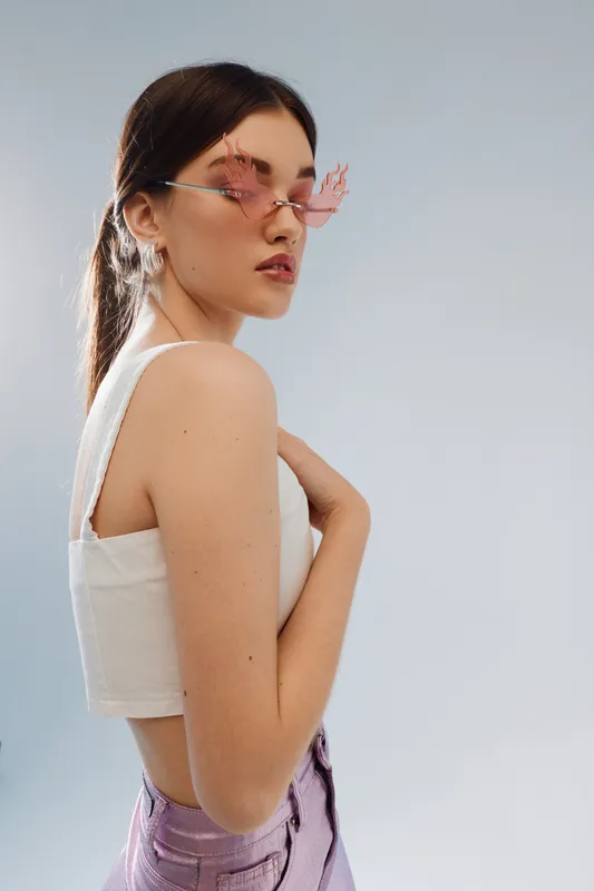 Young woman in stylish pink sunglasses and white top poses sideways against a light background. Concept of beauty, fashion, and modern style with confident expression.