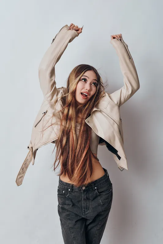 Young woman with long hair wearing beige jacket and black pants dancing joyfully with raised hands on light background. Casual style and expressive gesture with happy mood.