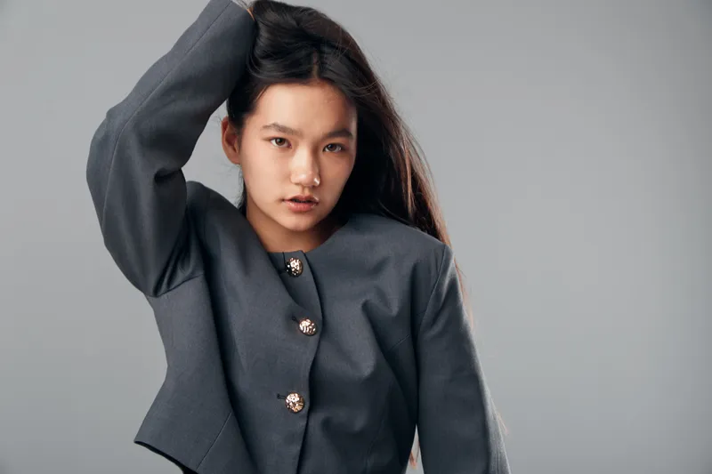 Woman with long hair in gray jacket posing with hand on head looking at camera in studio. Fashion portrait of female model with natural makeup on gray background and casual style.