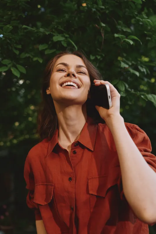 Young woman wearing a rust shirt smiles while talking on a phone in a lush leafy setting, creating a candid lifestyle moment outdoors
