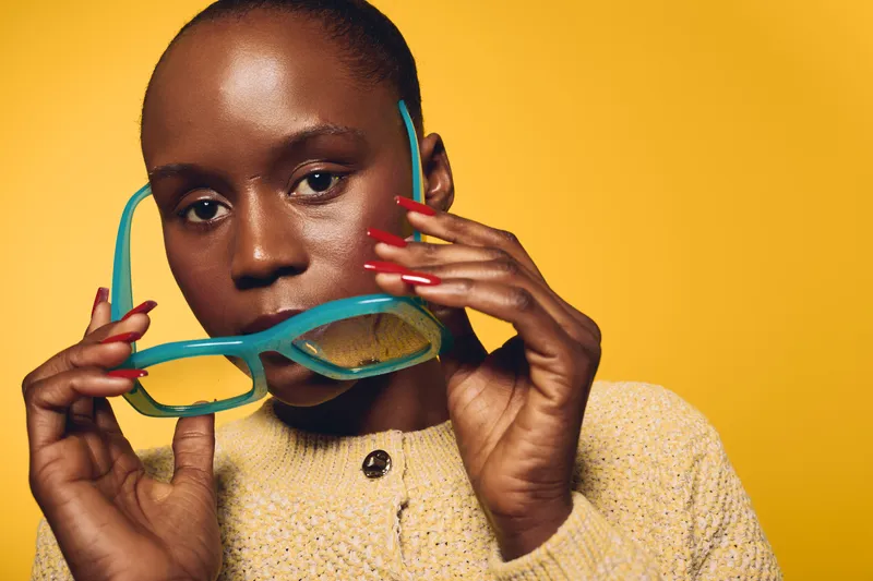 Close-up of dark skinned African woman holding turquoise glasses near face on yellow background. She wears beige knitted sweater with red nail polish. Concept of fashion, beauty, and style.