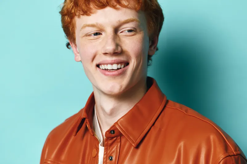 Redhead man with freckles smiling and looking aside wearing brown leather jacket on blue background. Young adult male with casual outfit in studio portrait.