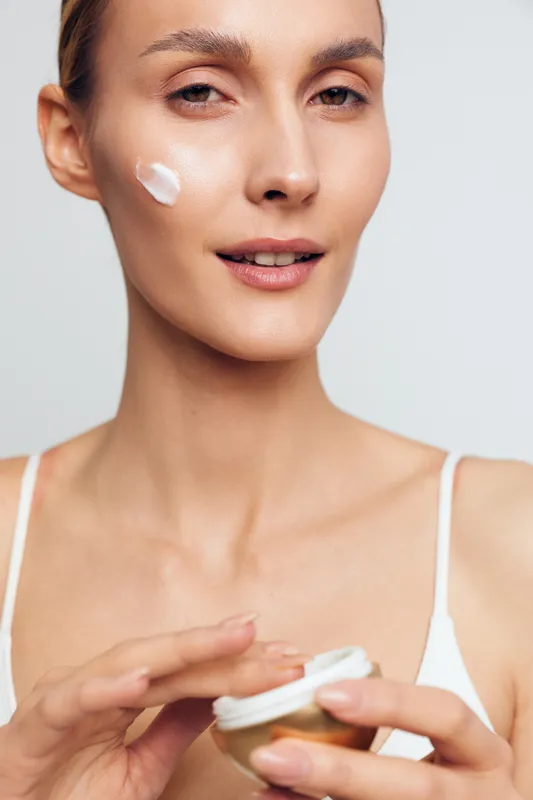 Woman applies face cream moisturizer on cheek, close up on clean skin against light background, skincare beauty hygiene treatment, wellness cosmetic concept daily routine