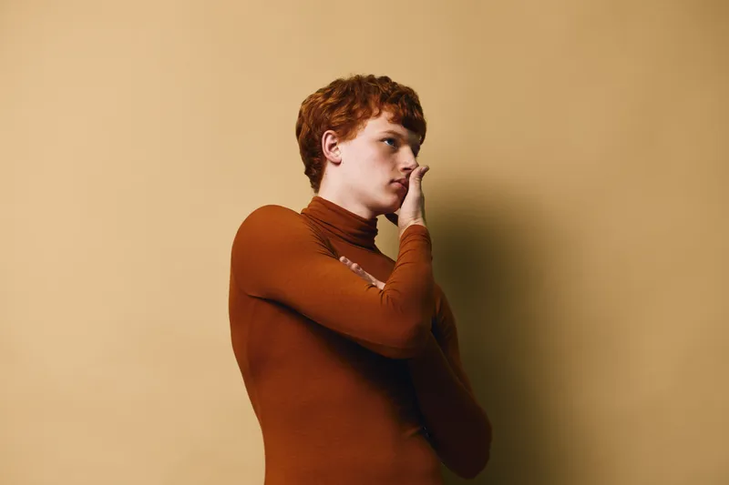 Young man with red hair wearing brown turtleneck sweater posing thoughtful with hand near face on beige background portrait in studio.
