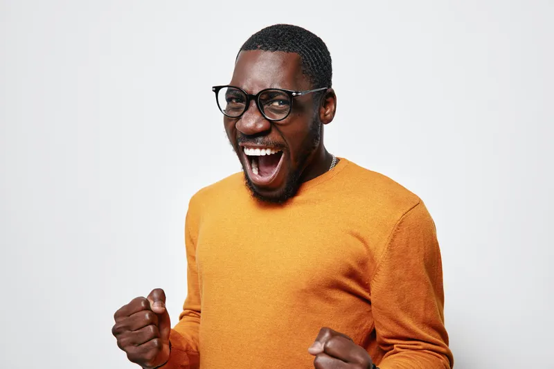 African American man with dark skin wearing glasses and orange sweater expressing excitement and joy with clenched fists on white background.