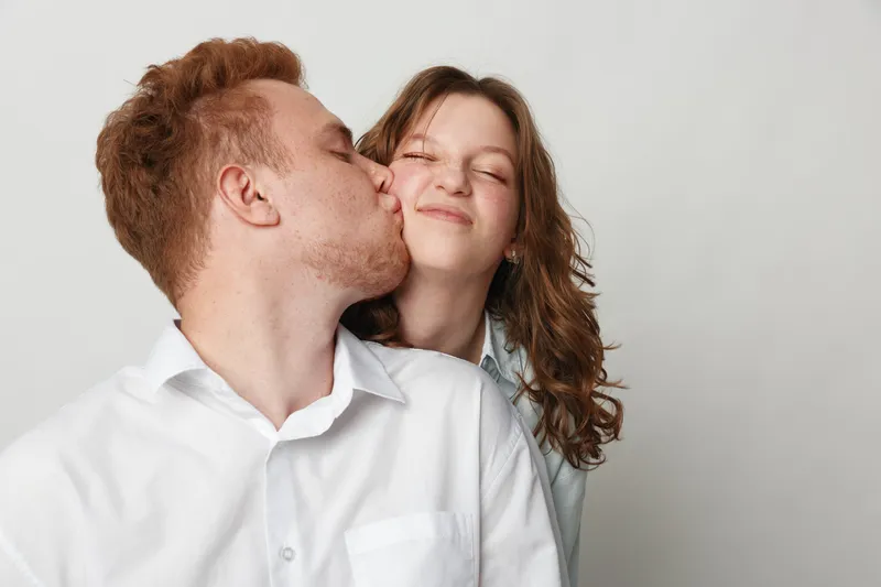 Young man kisses smiling woman on cheek in white shirts against plain background. Couple shows affection and happiness, concept of love, relationship, intimacy, and positive emotions.