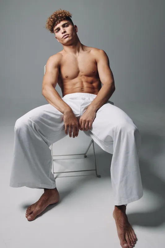 Young muscular man sitting on chair with bare torso and white loose pants in studio, posing confidently with relaxed posture and direct gaze at camera.