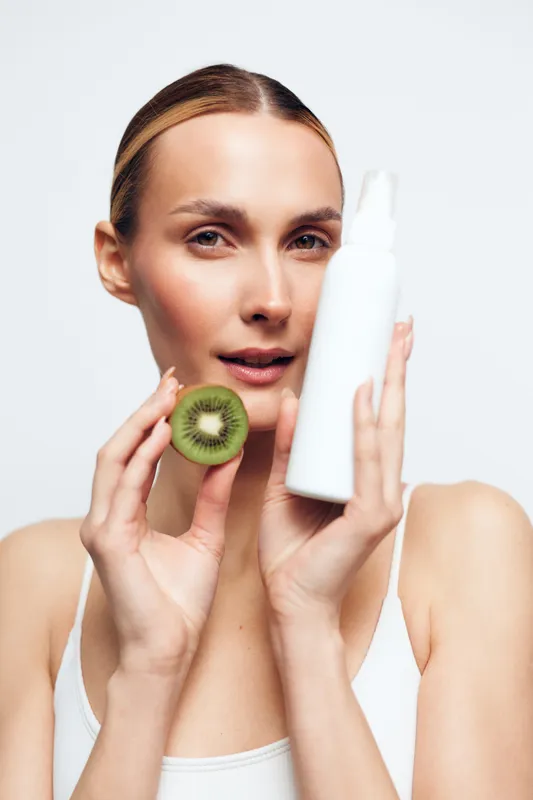 Young woman with clear skin holds half of fresh kiwi fruit and white cosmetic bottle close to face isolated on white background, beauty and skincare concept with natural ingredients.
