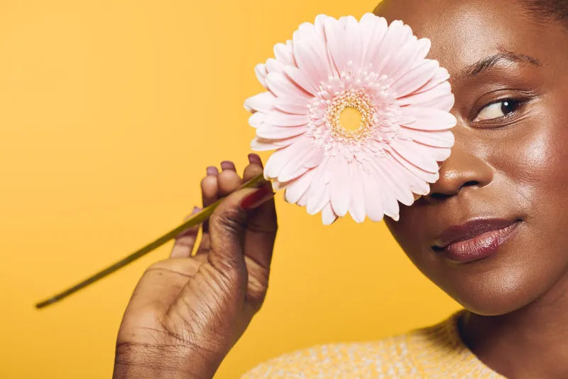 Close-up of a smiling dark skinned African woman holding a pink flower near her face against yellow background. Concept of beauty, nature, skincare, and happiness with natural light.