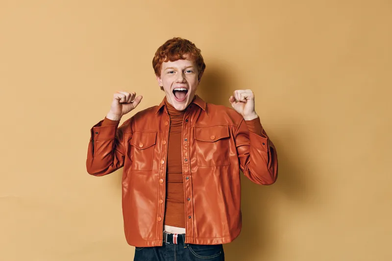 Young man with red hair celebrating success with clenched fists and open mouth, wearing brown leather jacket and turtleneck against a plain beige background.