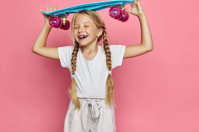 Happy young girl with braids holds a blue skateboard above her head with eyes closed. She smiles wearing a white t-shirt and light pants against a pink background.