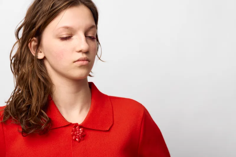 Young woman with long brown hair wearing red sweater looks down with serious expression on white background. Concept of contemplation, emotion, and casual fashion portrait.