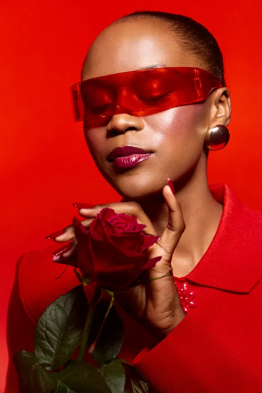 Confident black woman with dark skin wearing red sunglasses and red outfit holds a red rose near her face against red background. Concept of beauty, fashion, and elegance.