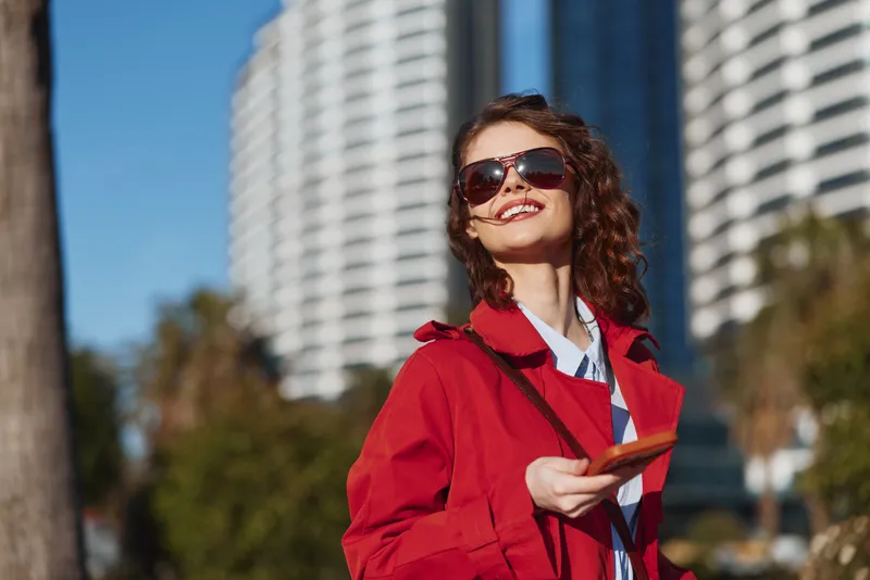 Smiling woman with curly hair wearing red coat and sunglasses holding smartphone outdoors in city. Bright daylight and modern buildings in background create lively mood.