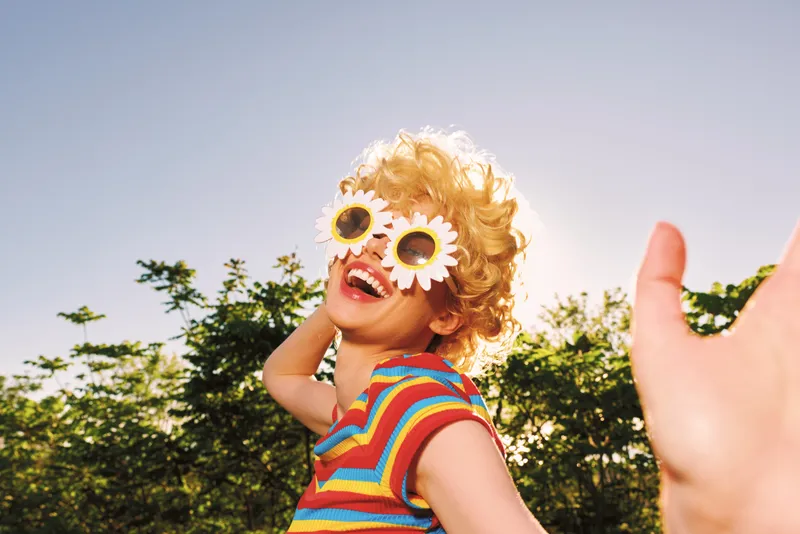 A young girl with blonde curls wears flower sunglasses and a colorful striped shirt, running outdoors under a bright sunny sky, capturing a playful retro moment.