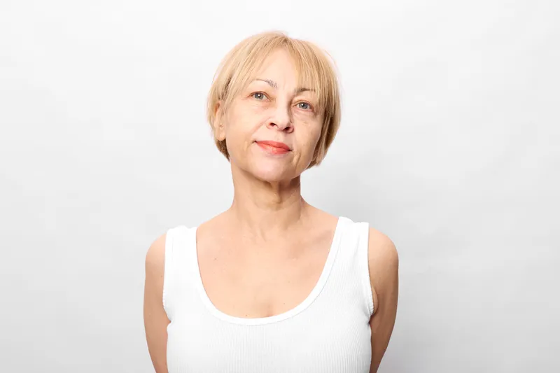 Smiling elderly woman portrait in studio light, looking at camera, wearing white tank top, neutral gray background, calm expression, soft focus, natural skin