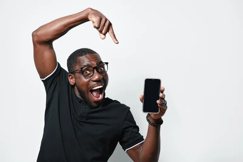 African American man with dark skin and glasses excitedly pointing at smartphone screen in black shirt on white background for technology and communication concept.