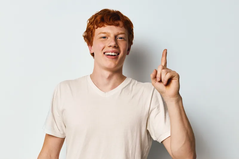 Young man with red hair smiling and pointing finger up on white background. Casual clothing, positive expression, standing and looking at camera with cheerful mood.