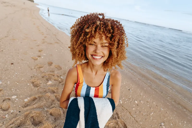 Woman on beach by ocean smiling holding towel, curly hair in sunlight, vacation mood, bright summer morning, relaxed posture, soft waves and sand texture, natural colors, outdoor leisure