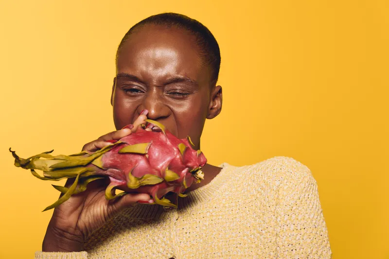 African woman with dark skin wearing beige sweater bites into fresh dragon fruit. Yellow background highlights natural texture and vivid colors. Concept for healthy eating and exotic fruit.