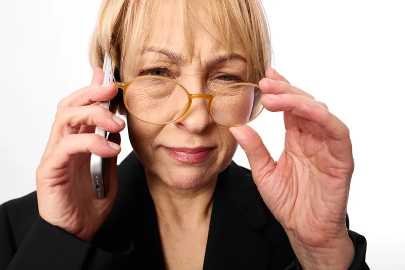 Elderly woman with glasses holding a phone to her ear, adjusting frames with both hands, calm confident portrait, focused look, white background, office style, closeup studio lighting.