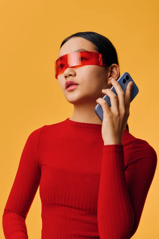 Woman in red sweater with futuristic red visor talking on smartphone on orange background. Modern style, casual pose, technology and communication concept with young female model.